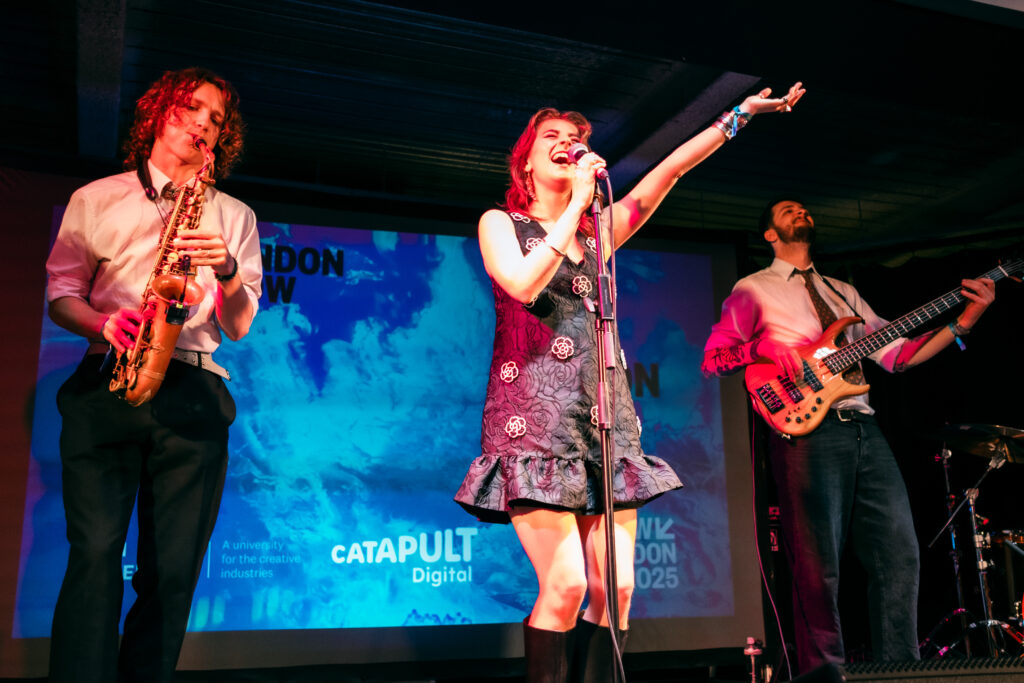 Female student singing into a microphone on stage, while two students play instruments next to her